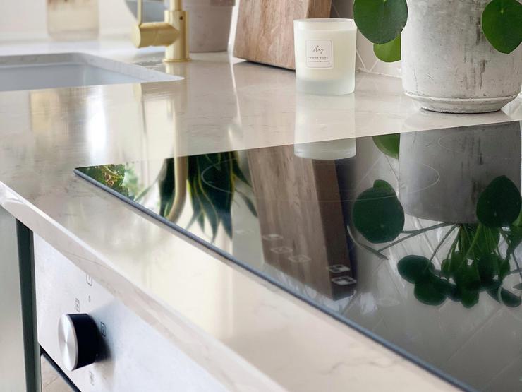 Close up of a white quartz worktop and a black induction hob, showing a brass mixer tap, white chevron tiles and a plant.