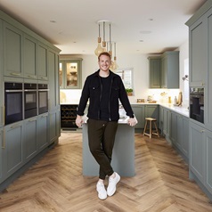 Greg Rutherford stood at the end of a kitchen island, showing sage green shaker cupboards and chevron wood flooring.