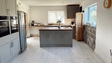 Old makeshift kitchen with an island, grey doors, light grey flooring, an exposed brick wall and rubble in doorway.