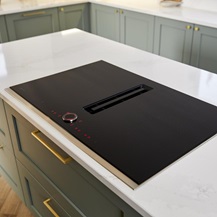 Black induction hob with a shiny surface on a reed green shaker island, with brass handles and a white quartz worktop.