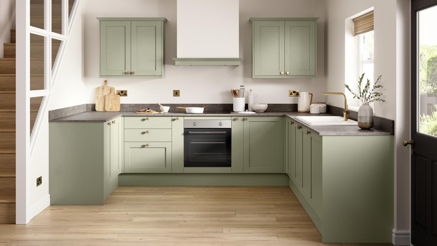 An l-shaped shaker kitchen in a pale, reed green colour. It has grey stone worktops and oak-style, laminate plank flooring. 