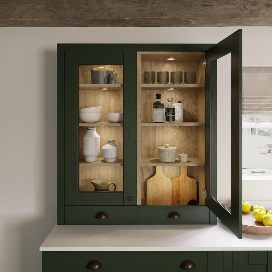 Dark-green kitchen cupboard with a glass door front, showing wooden shelves and neutral crockery, above a white worktop.