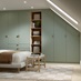 A modern bedroom with matt, reed green wardrobe doors and drawers. Behind the bed is dark wood panels and oak flooring.