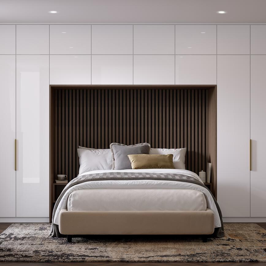 A bedroom with slab, white cabinets in a gloss finish. There is a drawer desser and tall units alongside and above a bed.